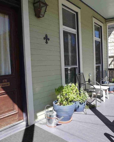 Covered porch with green siding, dark red wooden door, potted plants, and metal railing overlooking brick building