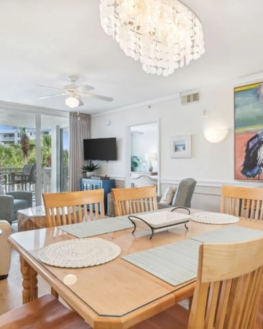 Bright open-plan living and dining area with wooden table, upholstered chairs, balcony view, and pelican artwork