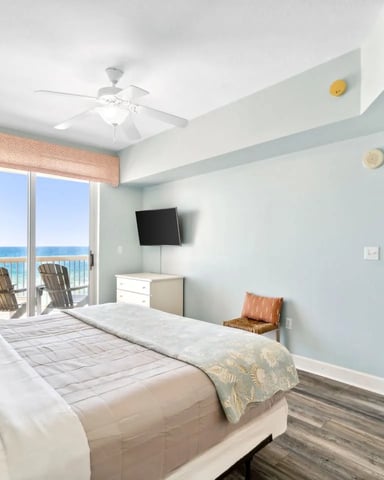 Beachfront bedroom with light blue walls, queen bed, wall-mounted TV, and ocean view from balcony with lounge chairs