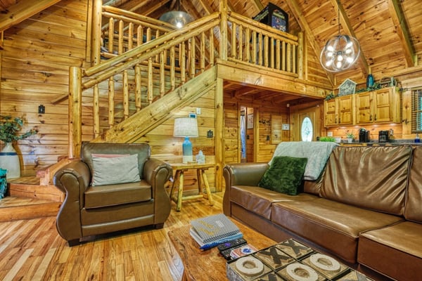 Log cabin living room with leather furniture, wooden loft staircase, and kitchen area visible in background