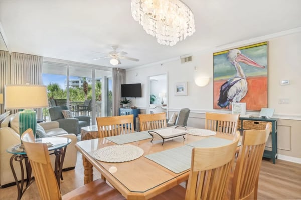 Bright open-plan living and dining area with wooden dining table, beige chairs, TV, and balcony overlooking palm trees